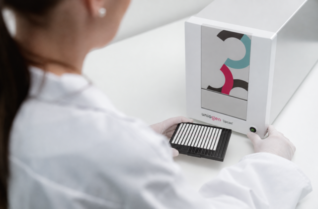Woman wearing a lab coat inserts a black lateral flow test strip holder with multiple white strips into an optical reader.